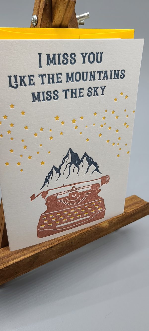 Letterpress printed A6 folding card. Says "I miss you like the mountains miss the sky" in dark blue. Has yellow stars below that, and a typewriter with mountainscape at the top. The card sits in front of a bright yellow envelope that matches the color of the stars on the card. Both the card and the envelope sit on a small, wooden easel.