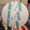 Photo shows a 6 inch diecut circle with the words FA LA LA printed in teal, down a row. There is a blind deboss to the left and right of the word, featuring the same text.