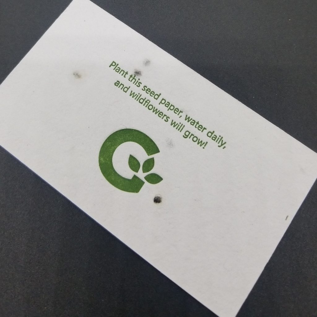 Photo shows a business card printed on handmade seed paper. The text says "Plant this seed paper, water daily, and wildflowers will grow!" Below that is the company logo, a large 'G' with three leaves coming out of the G.